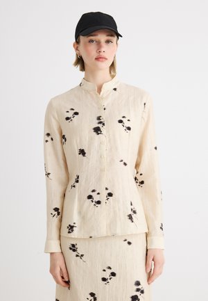 Frau trägt eine beige, langärmlige Bluse mit Knöpfen und passenden Rock mit schwarzem Blumenmuster, kombiniert mit einer schwarzen Baseballkappe.