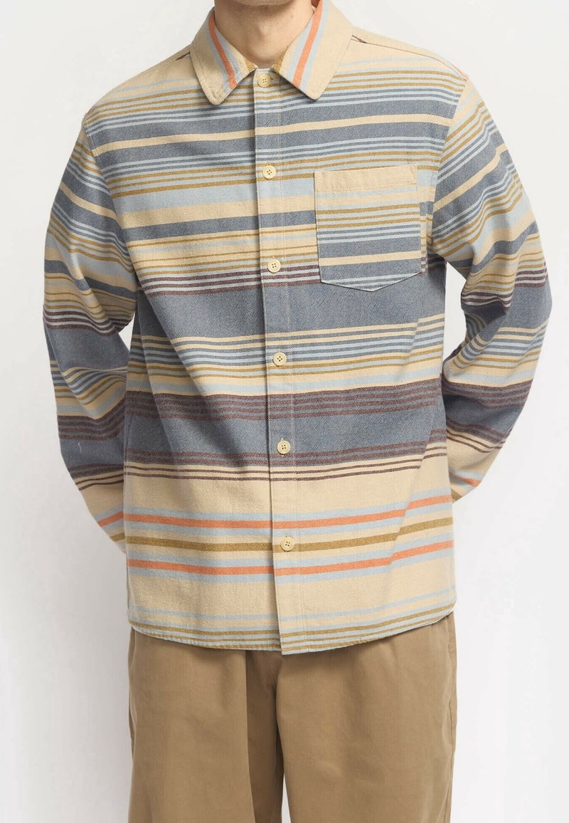 Homme portant une chemise à manches longues boutonnée avec des rayures horizontales bleues, beige, orange et marron, et un pantalon beige.