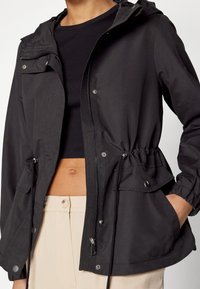 Giacca nera con cappuccio realizzata in materiale leggero, dotata di una zip frontale, vita con cordino e due tasche laterali con chiusura a scatto.