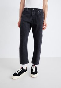 Jeans de mezclilla negra con un corte relajado, pierna recta y dobladillo deshilachado. Combinados con zapatillas negras con cordones blancos y suela color crema.