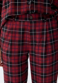 Un pantalon à carreaux rouges présente un motif en grille avec des lignes noires et blanches, une coupe ajustée avec un devant plissé et une fermeture à bouton.