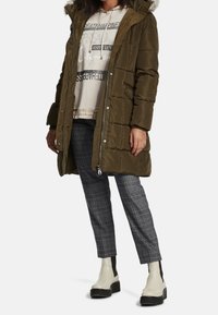 Kvinna i en olivgrön vadderad kappa med byxor i fuskpäls, grafisk beige huvtröja, grå rutiga byxor och krämfärgade plattformstövlar.