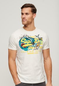 Camiseta de algodón blanca con estampado gráfico colorido de olas, una tabla de surf y texto. Diseño de mangas cortas y cuello redondo.