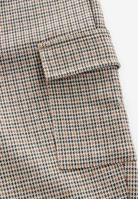 Pruun ja must houndstooth kangas, millel on väike tasku volditud klapiga, tekstuurne pind ja keerukad mustrid.