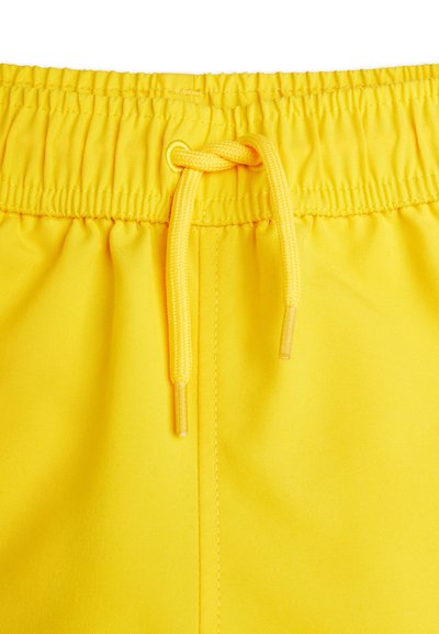 Ceinture élastique jaune avec un nœud de cordon de serrage noué sur un tissu léger, montrant les détails de couture et la texture.