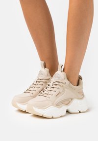 Baskets beiges avec une tige texturée, semelle blanche arrondie et design à lacets. Comprend des accents cousus et une languette.