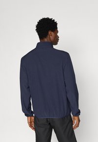 Sötétkék dzseki álló gallérral, rugalmas derékpánttal és hosszú ujjakkal. Simább anyag, minimalista dizájn, látható fémrészek vagy minták nélkül.