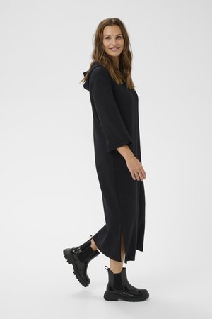 Une robe noire à manches longues en tissu doux avec une capuche et des fentes sur les côtés. Assortie à des bottes de cheville noires brillantes avec des côtés élastiques et une semelle épaisse.