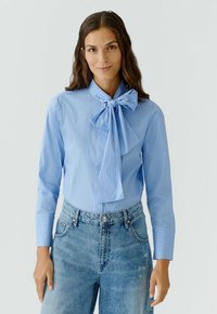 Lichtblauw gestreepte blouse met een grote strik bij de kraag, aansluitend in de taille, en lange mouwen, gecombineerd met high-waisted blauwe jeans.