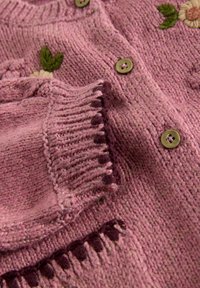 Vaaleanpunainen neulecardigan, jossa on vihreä lehti- ja keltakukkakirjonta, ruskeat napit ja taitettu hihansuu tummanruskeilla yksityiskohdilla.