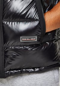 Gilet rembourré noir en matériau brillant, doté d'une fermeture éclair et d'un patch logo "KINGS WILL DREAM" à la taille. Texture lisse.