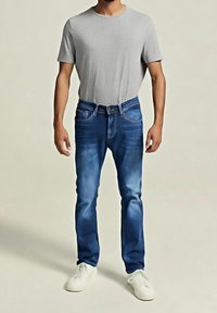 Mann trägt ein schlichtes graues T-Shirt, blaue Jeans und weiße Turnschuhe und steht vor einem neutralen Hintergrund.