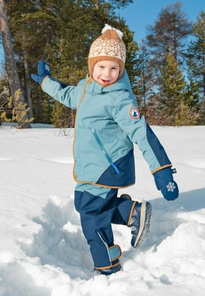 Giacca invernale blu e blu navy con dettagli arancioni, abbinata a pantaloni da neve e guanti coordinati. Cappello lavorato a maglia marrone con motivo bianco, sfondo innevato.