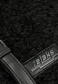 Černá chlupatá tkanina s texturovaným povrchem, doplněná černým koženým páskem a lesklou kovovou nášivkou s nápisem "relaks".