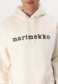 Krém színű kapucnis pulóver fekete "marimekko" felirattal, húzózsinórral ellátott kapucnival és puha textúrával, amelyet a ruhát viselő személy a mellkastól felfelé mutat.