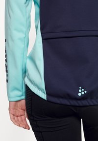 Chaqueta de forro polar con un diseño dividido en teal y navy, con un logo visible en la manga y acentos geométricos en la parte posterior.