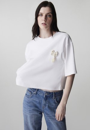 Mujer con cabello largo y oscuro, vistiendo una camiseta blanca recortada con un broche de lazo de cuentas y jeans de mezclilla azul, sobre un fondo liso.