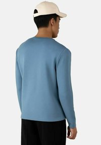 Lyseblå langermet genser med rund hals, teksturert stoff og minimalistisk design, båret med sorte bukser og en kremhvit caps.