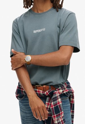 Person trägt ein graues Superdry-T-Shirt, kariertes Hemd um die Taille gebunden, blaue Jeans, braunen Gürtel und eine goldene Armbanduhr am linken Arm.