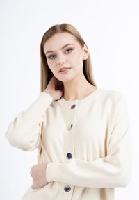 Cardigan lavorato a maglia color crema con scollatura rotonda, caratterizzato da bottoni scuri e polsini a coste. Tessuto morbido e testurizzato con una vestibilità rilassata.