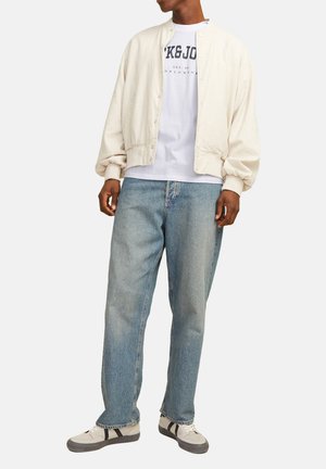 Helle beige, lässige Bomberjacke mit elastischen Bündchen und Druckknöpfen über einem weißen Hemd mit dunkelblauer Schrift. Getragen mit hellblauen Jeans und Sneakers.