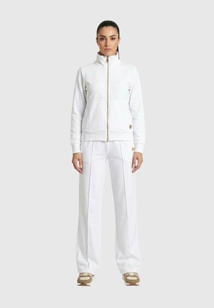 Une femme porte une veste blanche zippée et un pantalon assorti à jambes larges sur un fond uni clair.