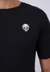 Black cotton t-shirt featuring a small white skull emblem on the left chest area. Short sleeves with a classic round neckline.