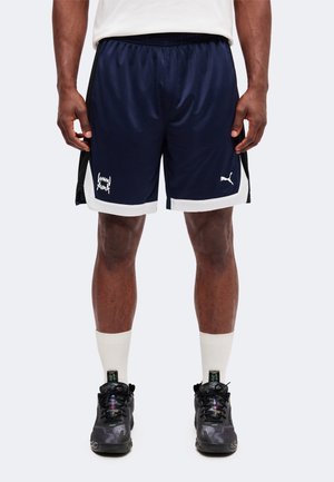 Homme portant un short de sport Puma bleu marine avec bordure blanche, des chaussettes blanches et des baskets noires, debout devant un fond clair uni.