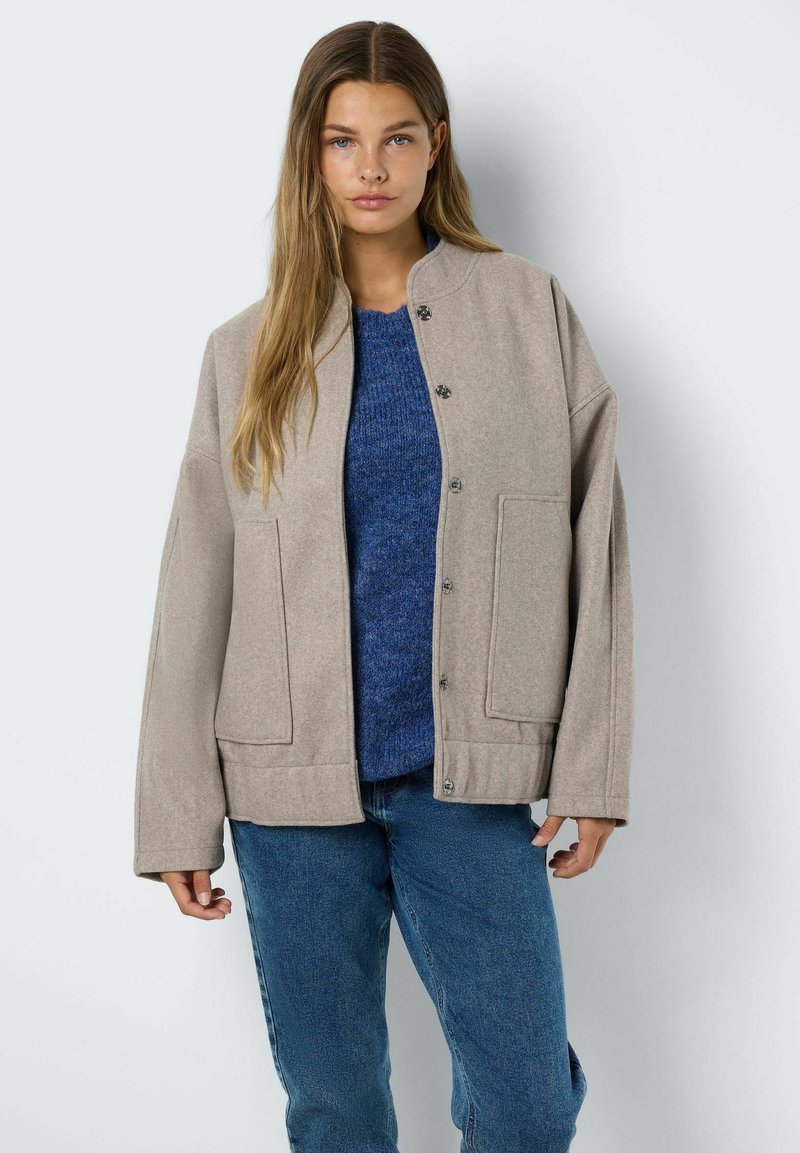 Veste en mélange de laine beige avec un col rond, des boutons sur le devant et deux grandes poches, portée sur un pull bleu et un jean.