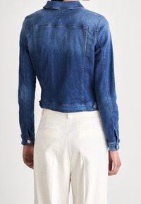 Jeansjacka i mediumblått med en figursydd design, kort längd och ärmar med knappar. Har klassiska axelsömmar och bakre ok.