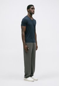 Άνδρας σε προφίλ ντυμένος με ναυτικό μπλε V-neck μπλουζάκι, χαλαρές γκρι παντελόνες και λευκά αθλητικά παπούτσια, σε απλό λευκό φόντο.