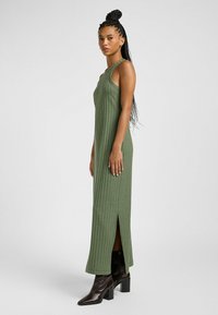 Abito maxi a coste verde con spacchi laterali, spalline larghe e silhouette aderente. Indossato con stivali neri alla caviglia, presenta un aspetto slanciato.