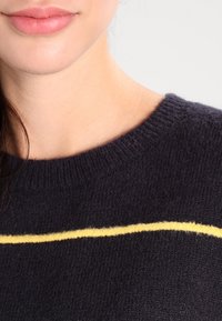 Marinblå stickad tröja med rund halsringning, med en framträdande gul rand över bröstet. Mjuk textur och ribbad krage.