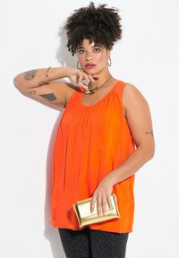 Top senza maniche di un vivace arancione con dettagli plissettati, abbinato a leggings neri con motivo e una piccola pochette dorata. Texture del tessuto liscia.