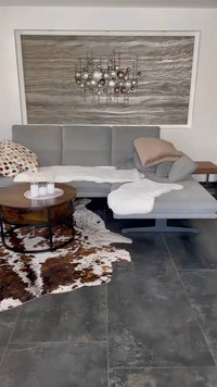 Harmaa kulmasohva pehmeällä heittotyynyllä, pyöreä puinen kahviteline ja lehmännahkamatto. Seinällä on abstraktia metallikoristetta. Tumma laatoitettu lattia.