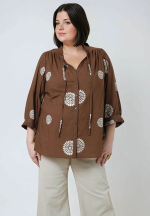 Brown blouse with floral embroidery, button front, gathered neckline, and 3/4 sleeves with ruffle detail. Paired with light-colored pants.