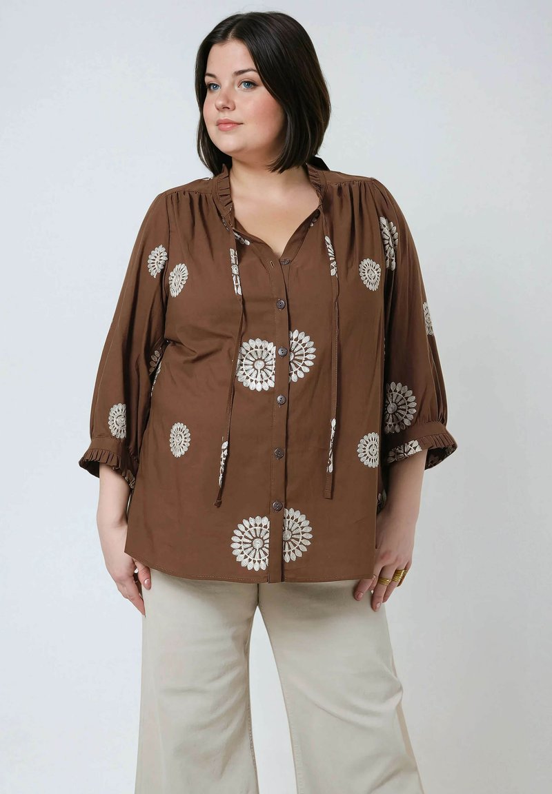 Bruine blouse met bloemenborduursel, knoopsluiting aan de voorkant, geplooide halslijn en driekwartmouwen met ruches. Gecombineerd met lichtgekleurde broek.
