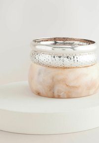Des bracelets métalliques empilés sur une base en marbre rose pâle lisse, présentant un design texturé et une finition brillante. Exposés sur une plateforme blanche.