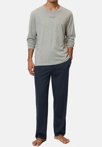 Svijetlosiva majica s dugim rukavima, okruglim izrezom i logotipom na prsima, u kombinaciji s tamnoplave široke hlače od mekog materijala.