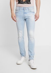 Jeans in denim blu chiaro con vestibilità slim, caratterizzati da dettagli strappati, un design a cinque tasche e chiusura con bottoni. Indossati con sneakers bianche.