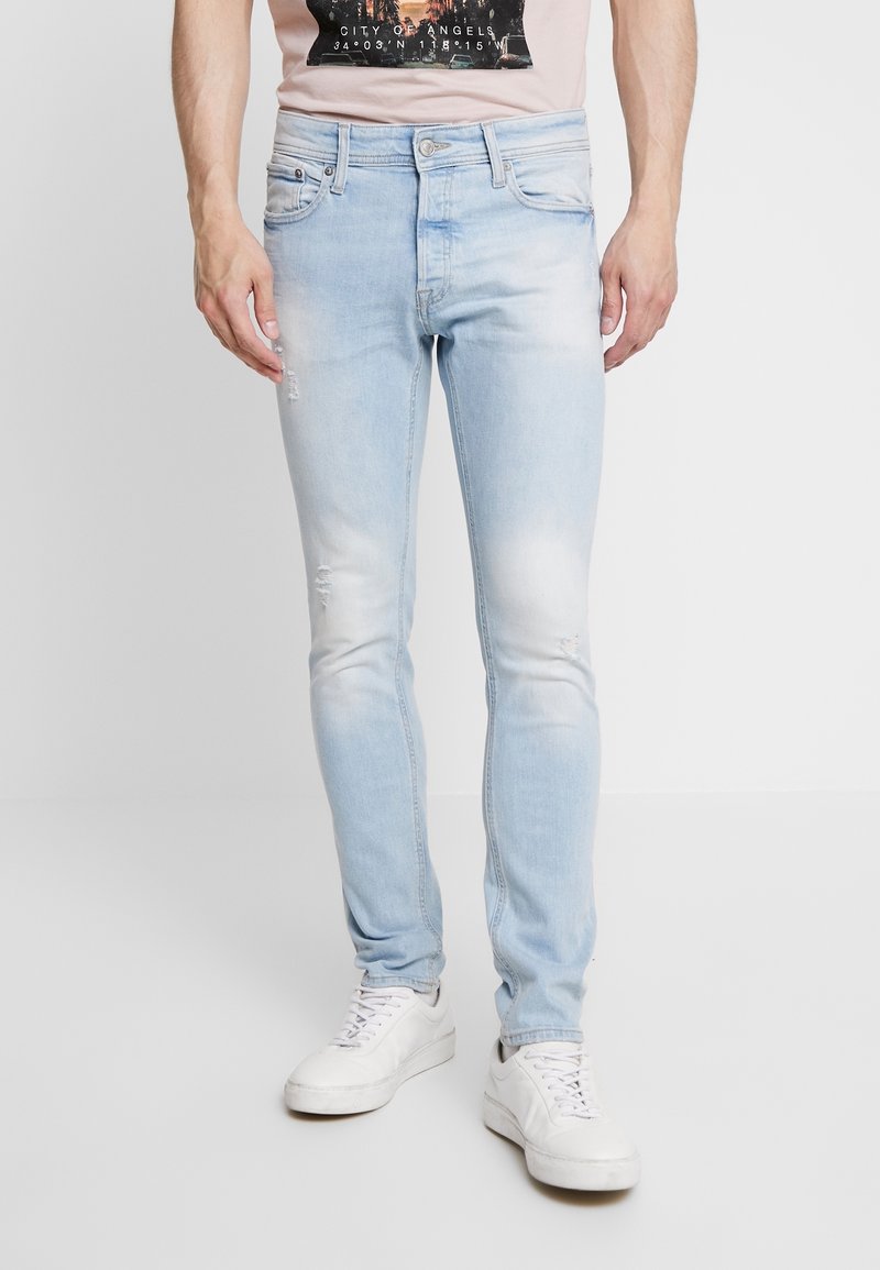 Jeans in denim blu chiaro con vestibilità slim, caratterizzati da dettagli strappati, un design a cinque tasche e chiusura con bottoni. Indossati con sneakers bianche.
