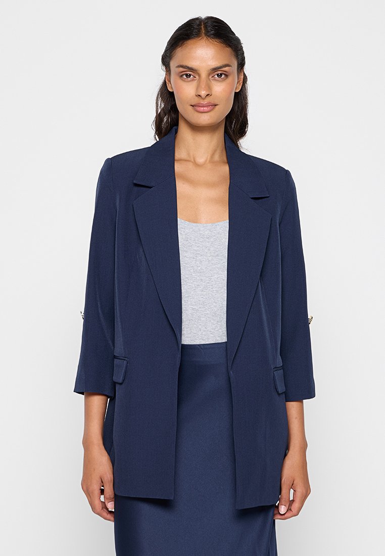 Blazer azul marino con mangas largas, solapas amplias y dos bolsillos frontales. Posee una textura suave y un ajuste ligeramente holgado.
