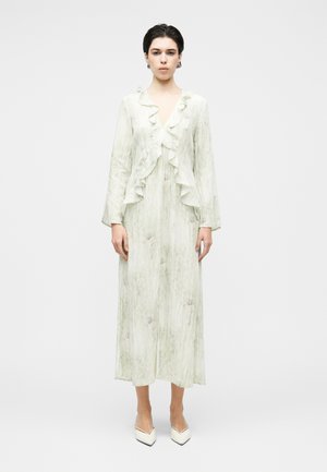 Młoda kobieta w długiej, jasnozielonej sukience z falbanami na dekolcie w kształcie litery V oraz długimi rękawami, w połączeniu z białymi szpiczastymi szpilkami.