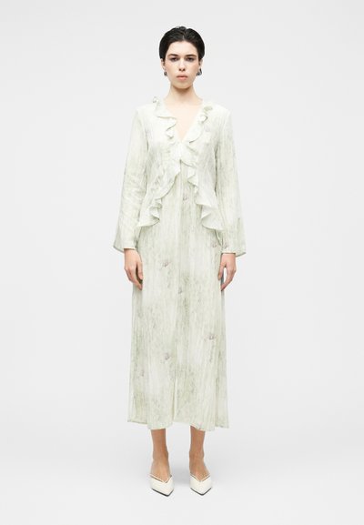 Jeune femme portant une longue robe vert pâle à encolure en V volantée et manches longues, associée à des escarpins blancs à bouts pointus.