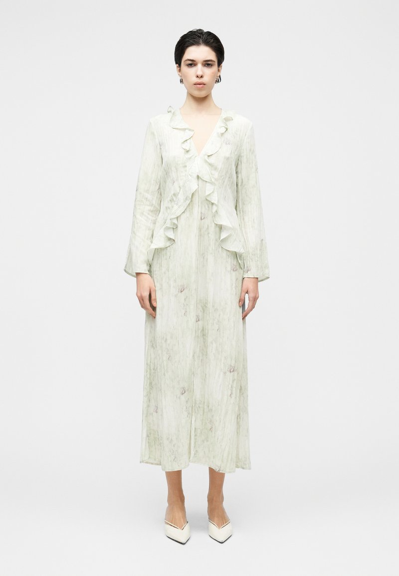 Νεαρή γυναίκα που φοράει μακρύ ανοιχτό πράσινο φόρεμα με βολάν στη V-λαιμόκοψη και μακριά μανίκια, σε συνδυασμό με λευκές μυτερές γόβες.