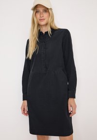 Schwarzes, langärmliges Hemdkleid mit Knopfleiste, seitlichen Taschen und weitem Schnitt, gefertigt aus einem weichen Stoff mit glatter Textur.