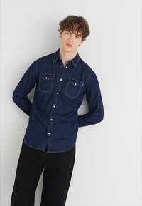 Donkerblauwe denim overhemd met lange mouwen, twee borstzakken, knopenlijst en contrasterende stiksels, gecombineerd met zwarte broek.