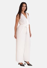 Hvid plisseret jumpsuit med dyb V-hals og bælte; ærmeløs design med svajende brede ben, har en glat stoftekstur.