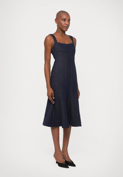 Námornícky denimová šatová bezrukávová šaty so štvorcovým výstrihom, siluetou A-line a švovými detailmi; doplnené čiernymi topánkami s ostrou špičkou.