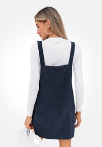 Vestido de pana azul marino con tirantes anchos, tejido texturizado y un corte holgado, combinado con una blusa blanca de manga larga y un bolso blanco.